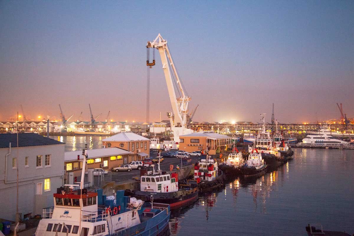 Cape Town harbour at twilight showing working crane, tugboats and industrial waterfront with illuminated facilities